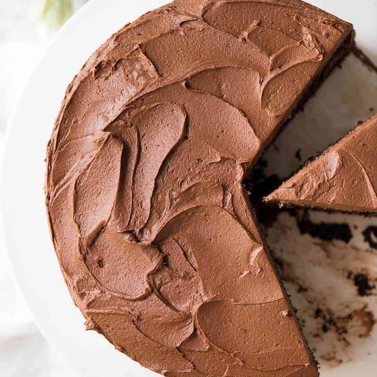 Chocolate Cake with Chocolate Buttercream Frosting on a white cake platter with a piece cut out, photographed from overhead.