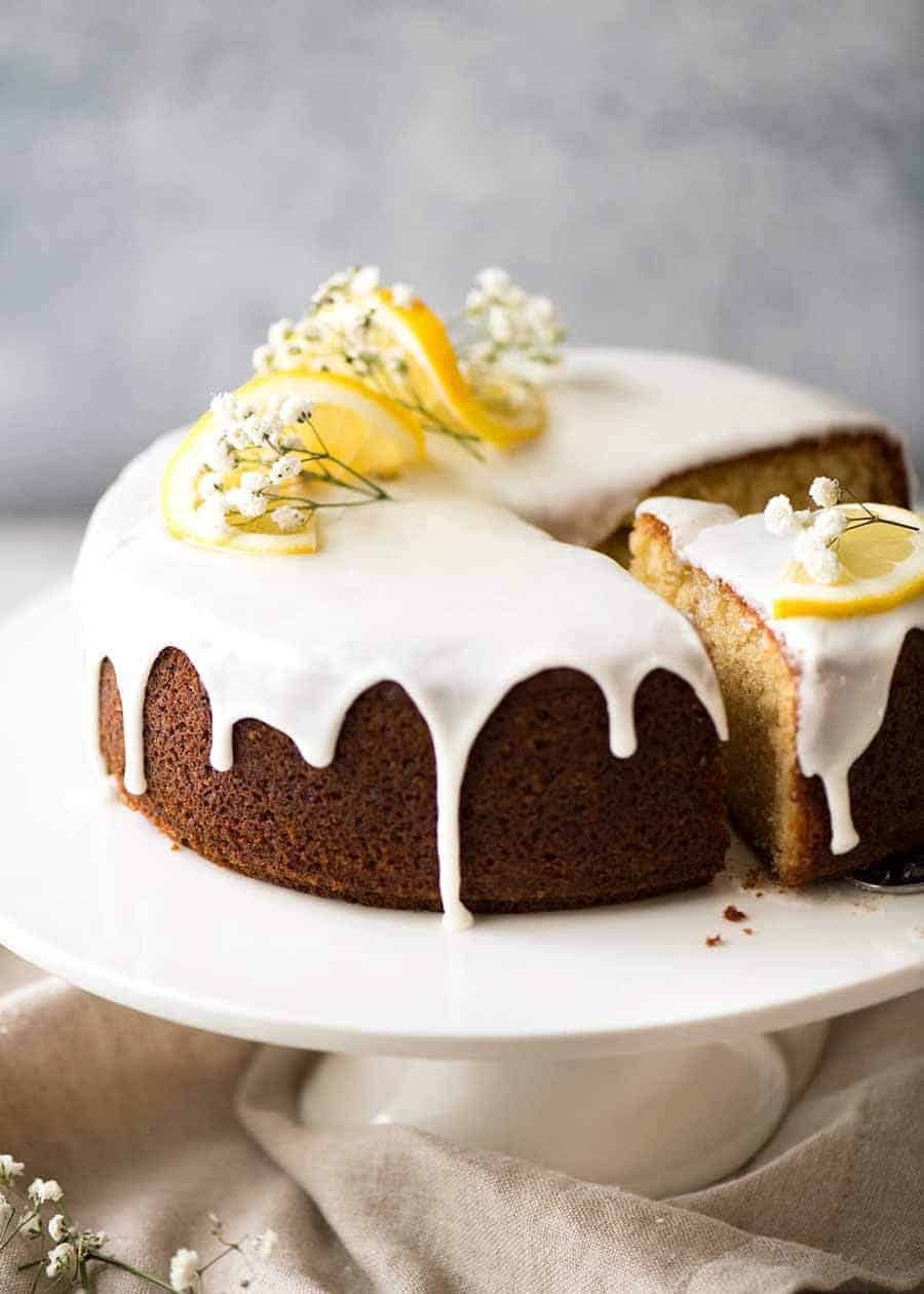 Lemon Cake with drippy lemon glaze decorated with lemon slices and flowers, on a white cake platter with a piece cut out