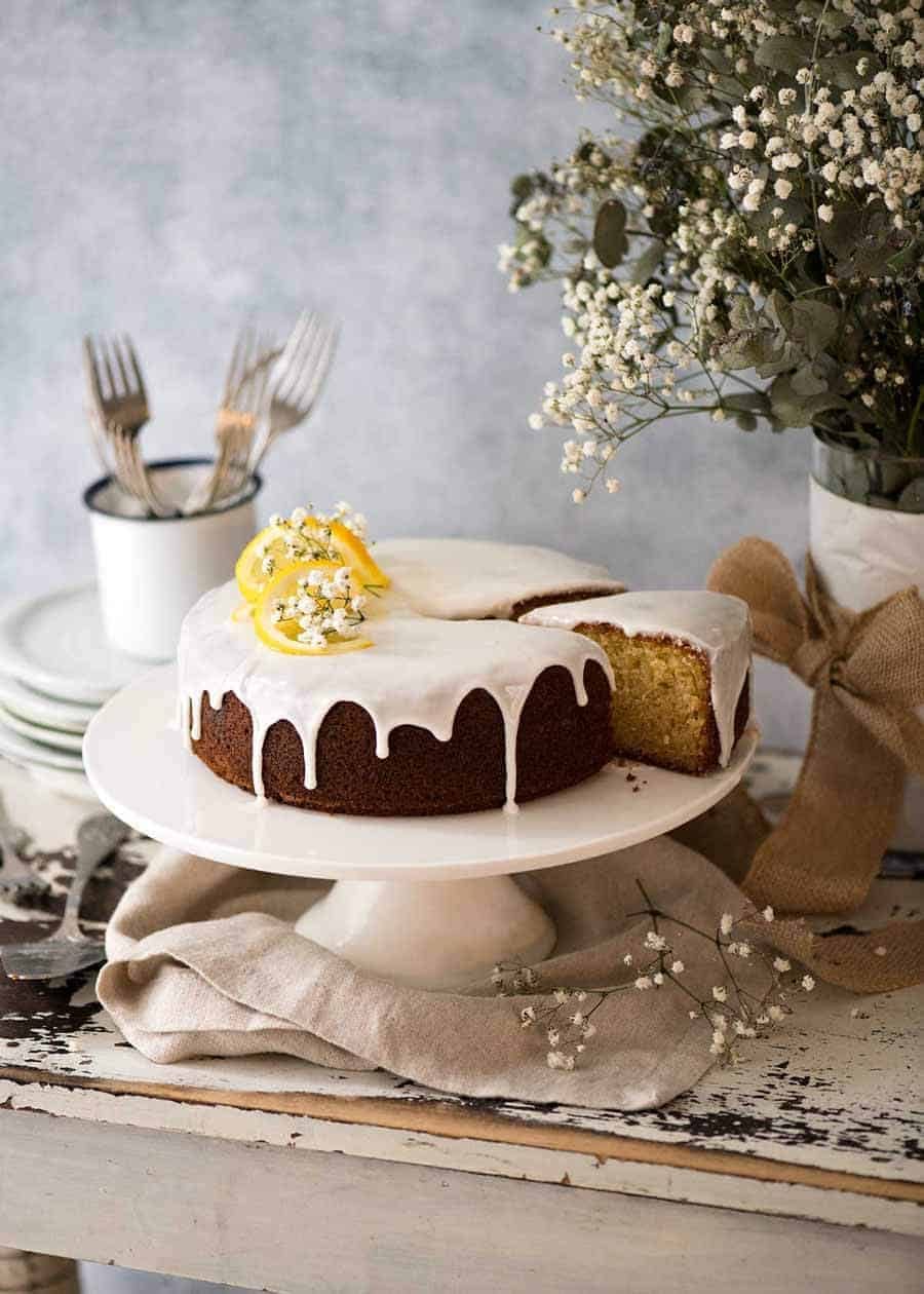 Lemon Cake with drippy lemon glaze on a white cake platter with a piece cut out and plates on the side, ready to the served.